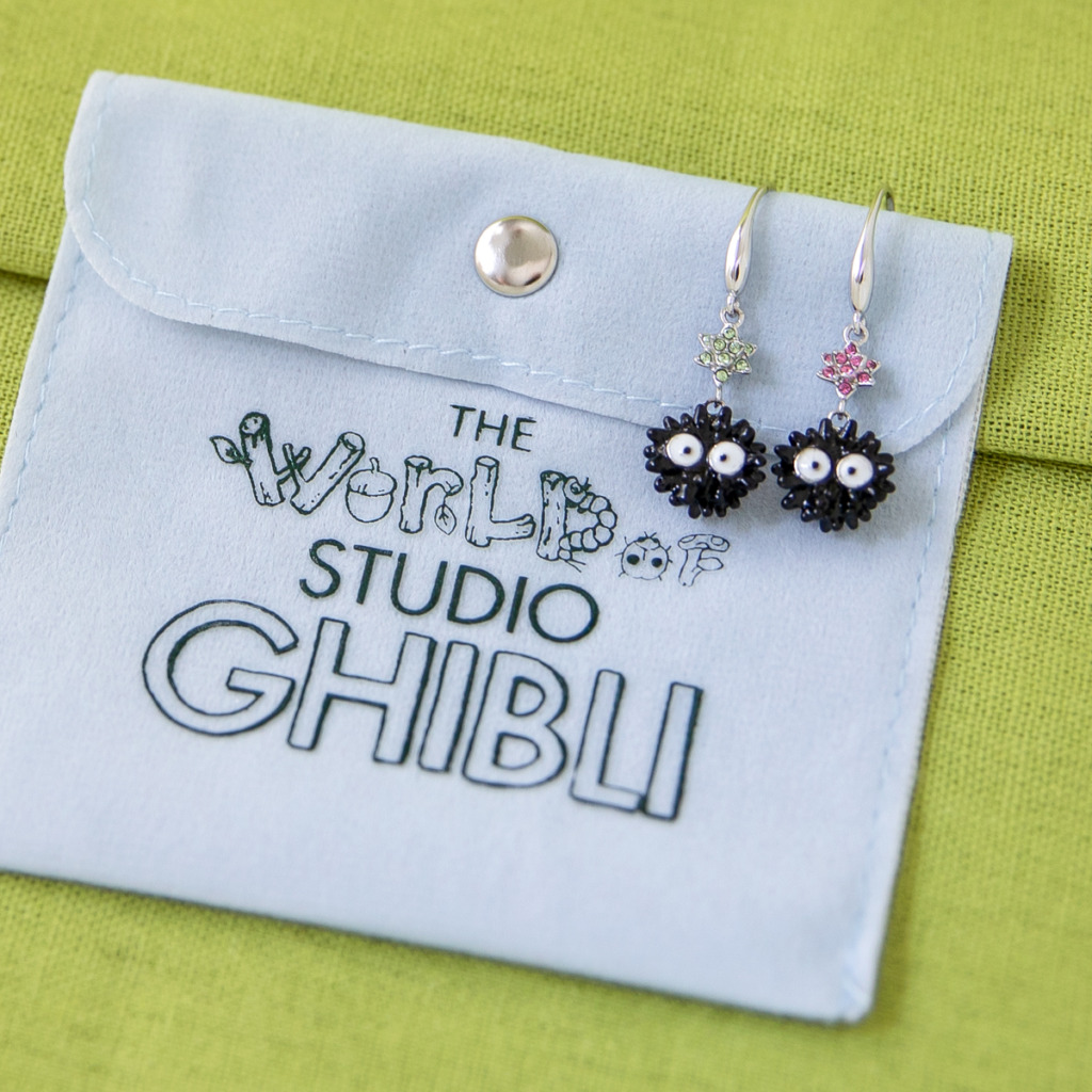 A pair of Soot Sprite earrings lays on top of a white travel pouch with The World of Studio Ghibli logo on it.