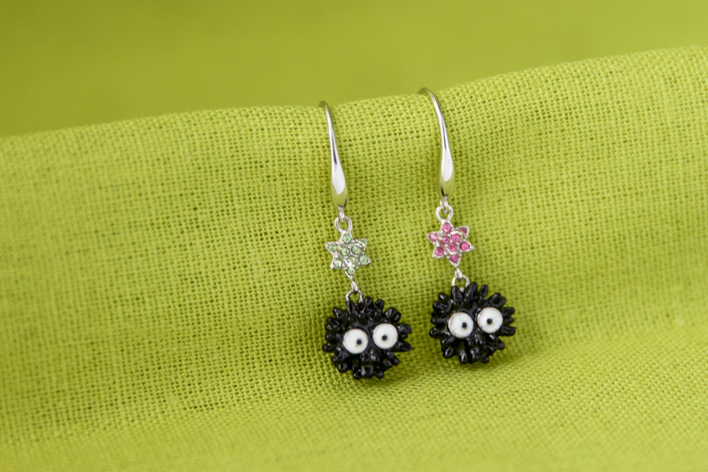 A pair of Soot Sprite Earrings with black enamel Soot Sprites and pave crystal candy stars against a green background.