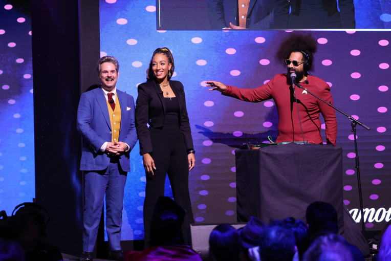 From Left to Right: Paul F. Tompkins, Tawny Newsome, standing side by side with Reggie Watts behind a DJ table stand at the Star Trek Day events 