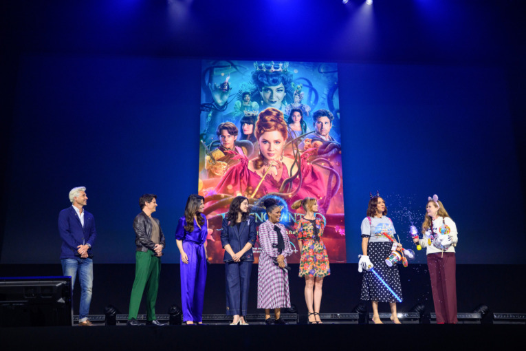 the cast of Disenchanted with the poster for the new movie displayed behind them at D23 Expo