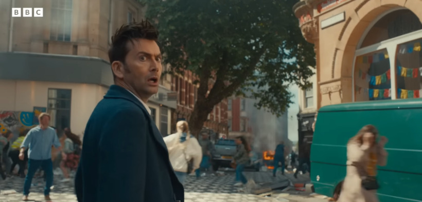 The 14th Doctor, played by David Tennant, looks behind him as people run in fear from something down the street