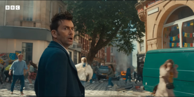 The 14th Doctor, played by David Tennant, looks behind him as people run in fear from something down the street