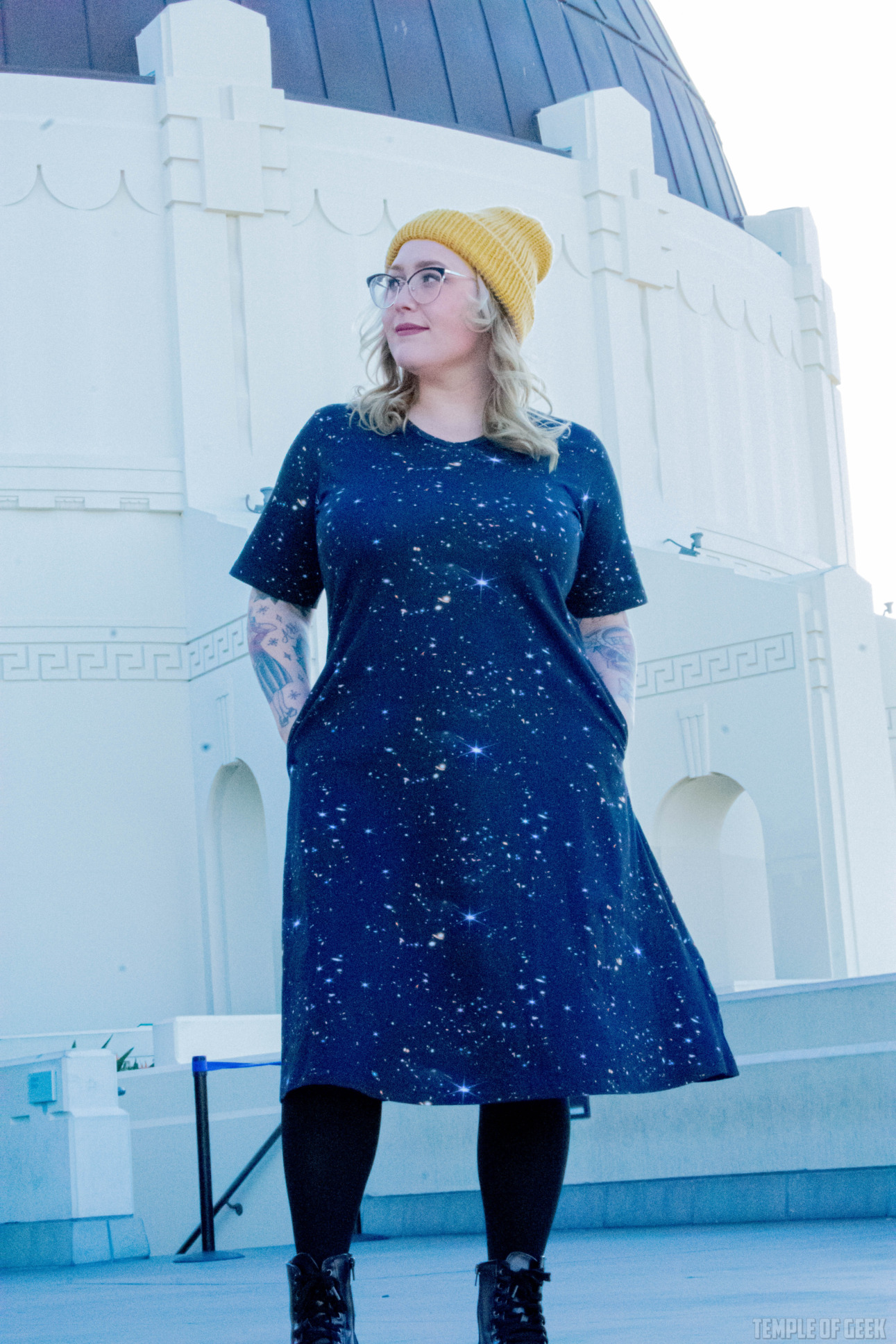 A model wearing a black dress with a space-themed pattern on it and a yellow beanie poses outside Griffith Observatory.