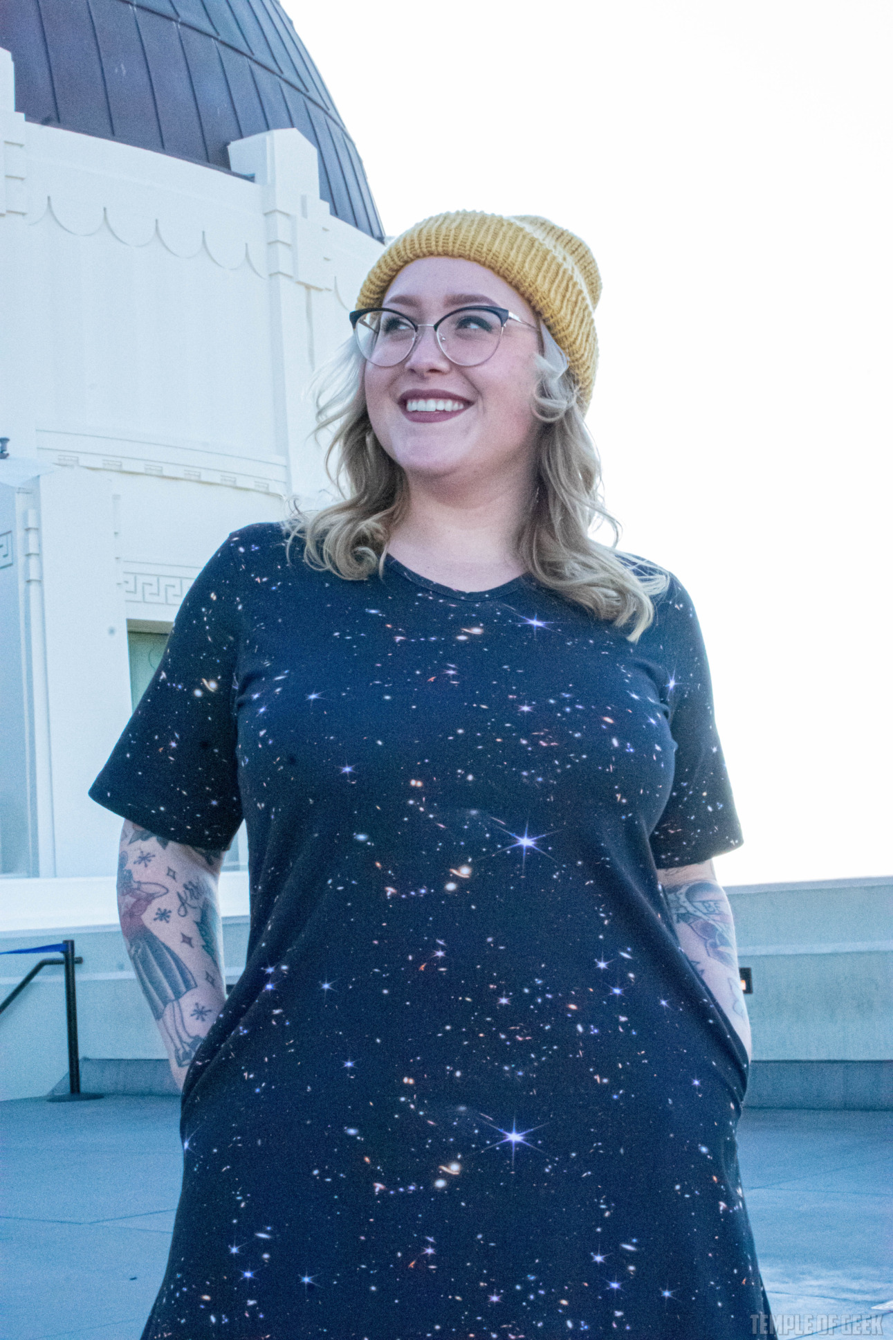 A model wears a yellow beanie and has her hands in the pockets of a black dress from Svaha outside Griffith Observatory.