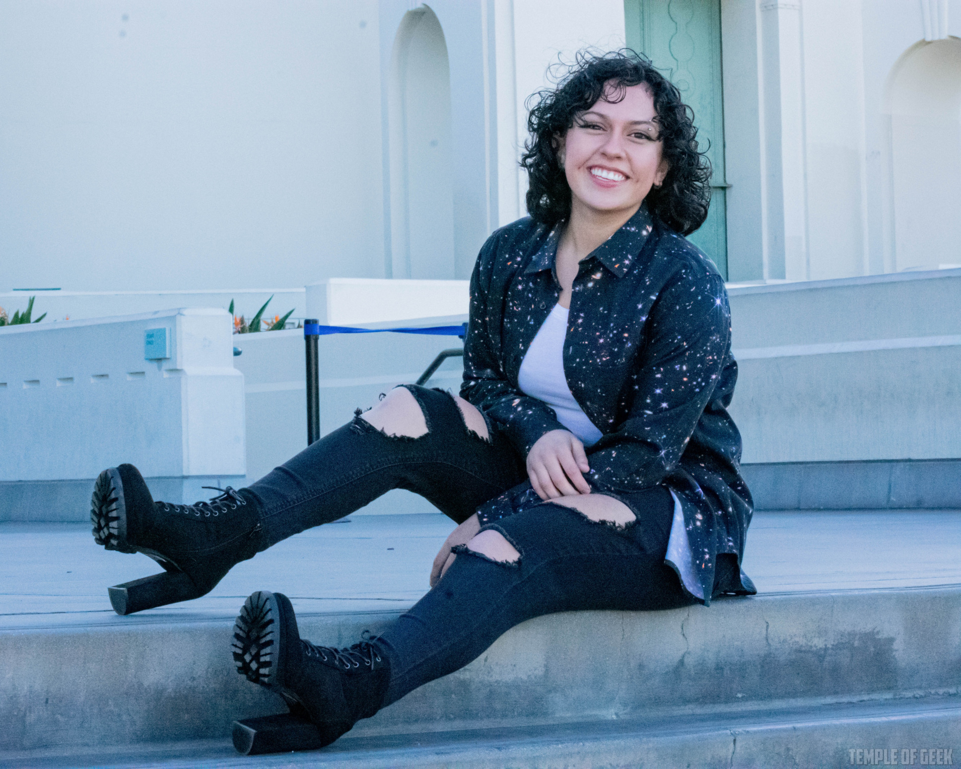 A model poses with her legs stretched out on steps at Griffith Observatory. She wears a black button-up from Svaha over a white tanktop and black jeans with high-heeled boots.