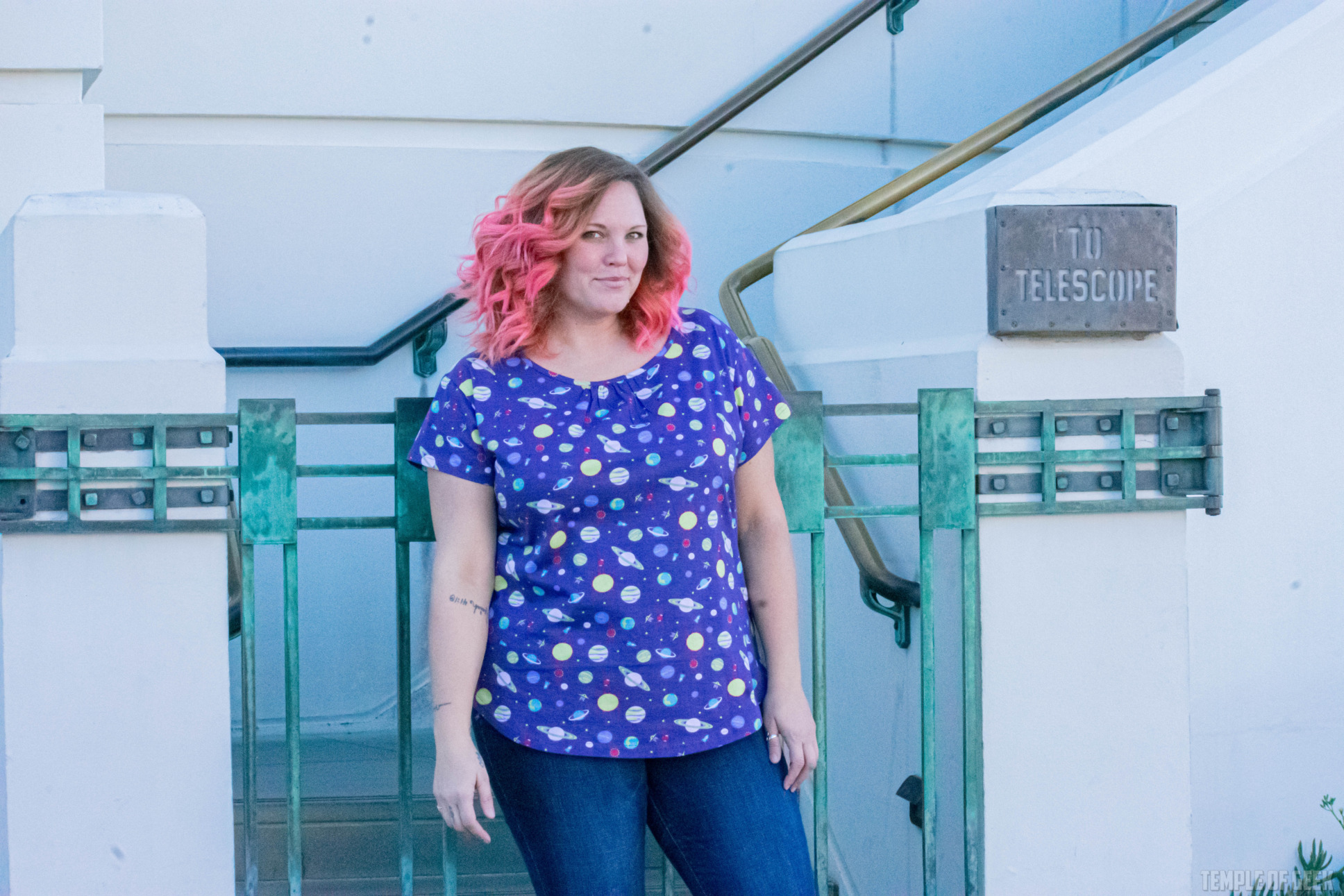 A model with pink hair poses against a gate at Griffith Observatory. She wears a space-themed top from Svaha.