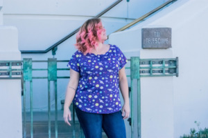 A model looks up white standing in front a gate that leads to a telescope at Griffith Observatory. She has pink hair and wears a purple, planet-themed top. 