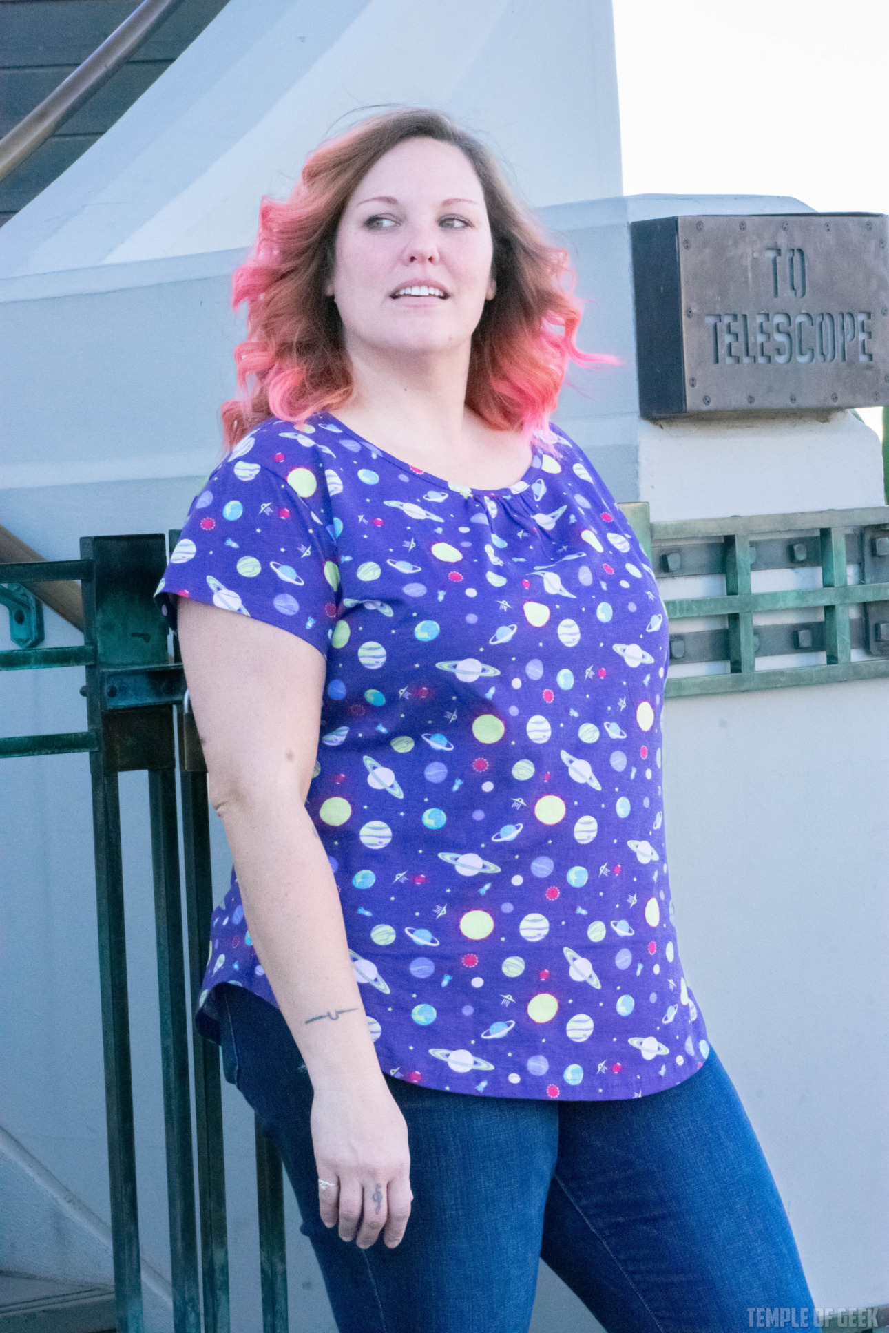 A model poses against a gate at Griffith Observatory. She wears a purple, short-sleeve top covered in little planets and other space-themed icons.