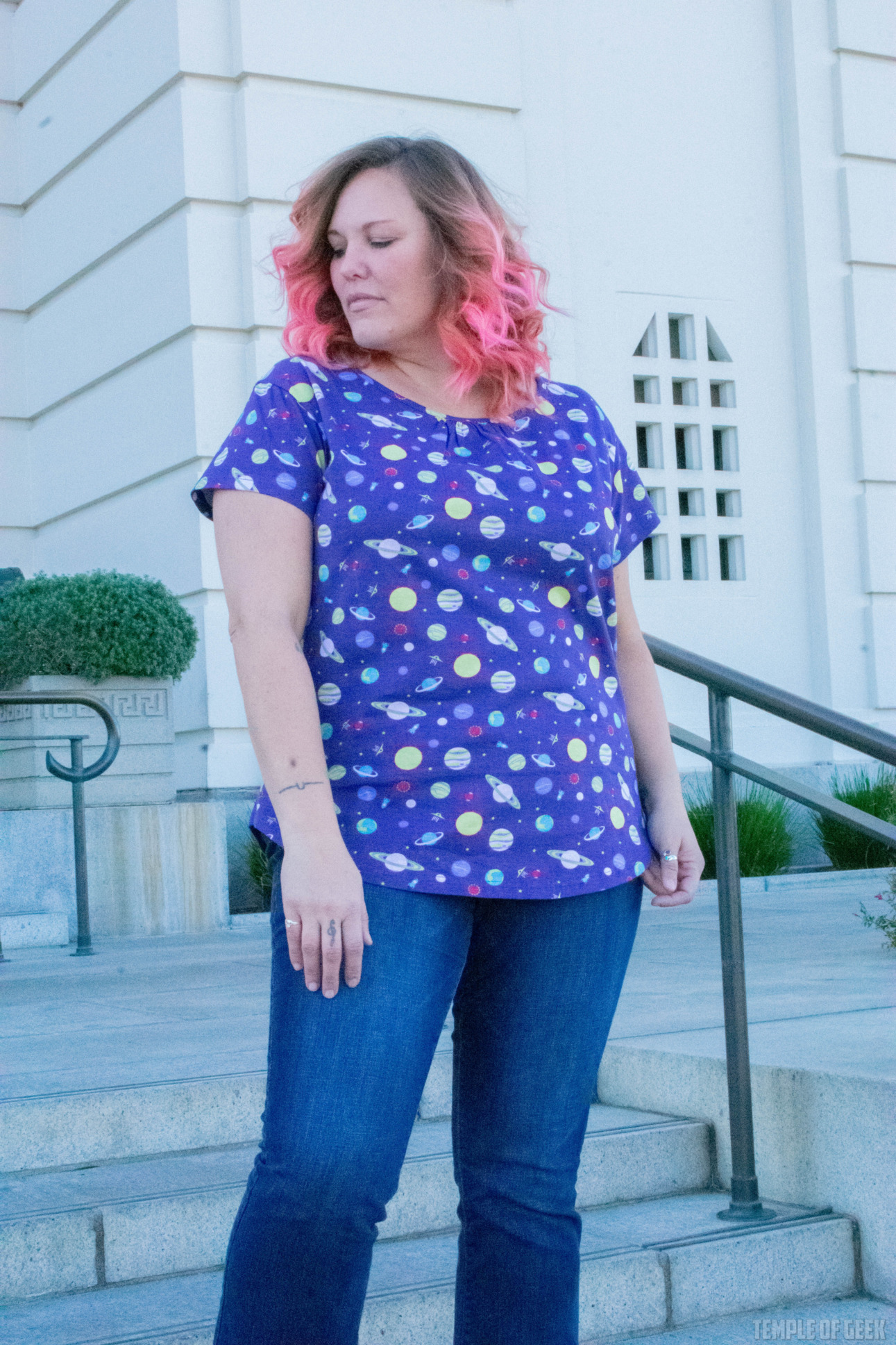 A model poses in front of a staircase at Griffith Observatory. She has pink hair and wears a purple, planet-themed top with blue jeans.