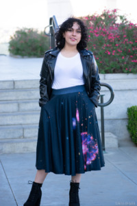 A model poses in front of a staircase with flowers next to it. She wears a leather jacket, white tee, and a space-theme twirl skirt from Svaha. 