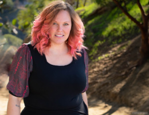 A model with pink hair stands in a park, wearing a black shirt with sheer puff sleeves with a gamer-themed print on them. 