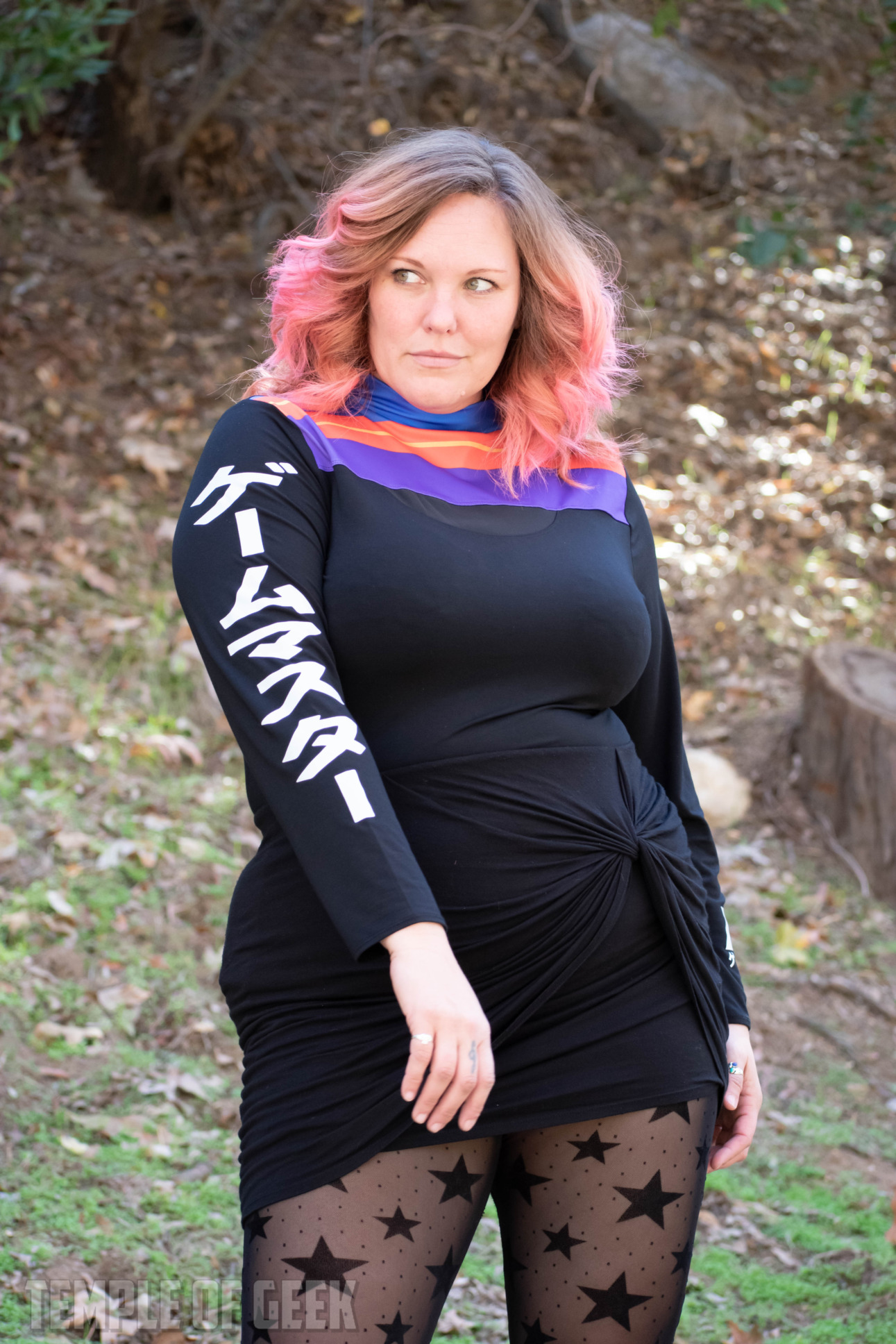 A model poses in a park wearing gamer fashion, consisting of a mock turtleneck top with stripes and Japanese characters and a black skirt with star-patterned black tights.