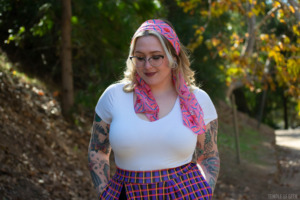 A model wears a pink, gamer-themed scarf in her hair, a white t-shirt, and a plaid skort. Her hands are in her pockets and she stands in a tree-filled park. 