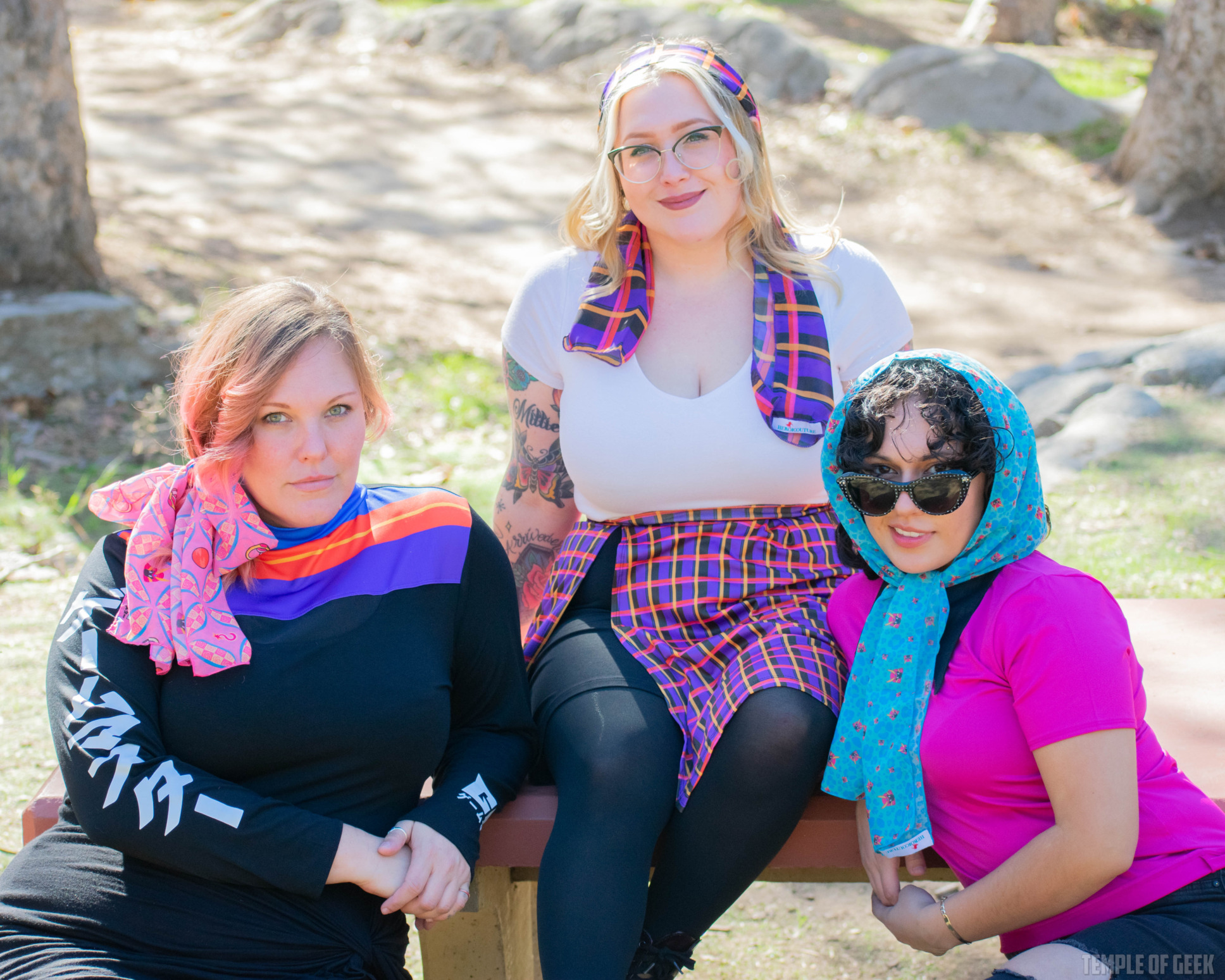 Three models sit at a picnic table in the park wearing brightly colored scarves from gamer fashion line GameMaster + Heroicouture.