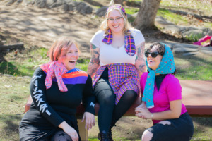 Three models laugh together while sitting at a picnic table in the park. They wear scarves in their and gamer fashion from GameMaster + Heroicouture. 