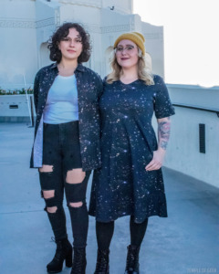 Two models stand outside Griffith Observatory. One wears a button-up shirt and one wears a fit and flare dress. The space-themed clothes are from Svaha and have a pattern from the James Webb Telescope on them. 