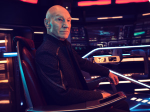 Patrick Stewart as Jean-Luc Picard sits in a chair surrounded by starship displays, controls and panels