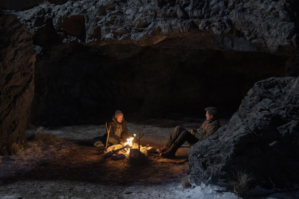 Ellie (Bella Ramsey) and Joel (Pedro Pascal) sit by a camp fire at night.