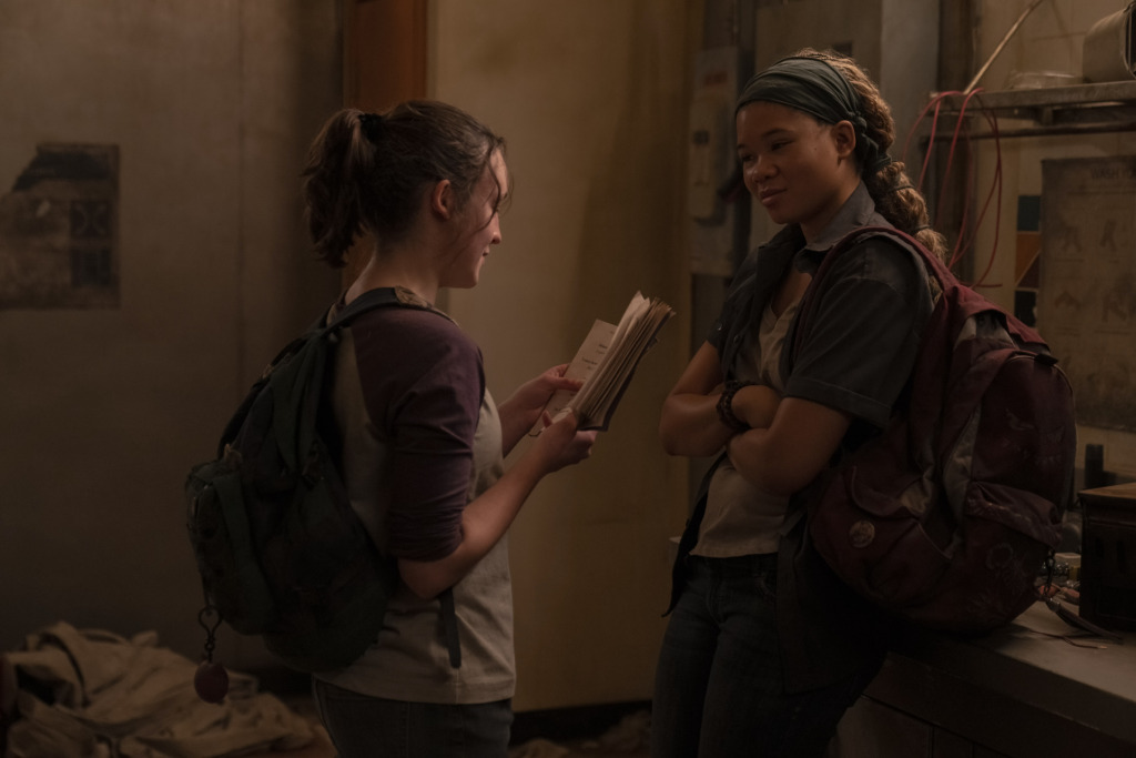 Ellie (Bella Ramsey) and Riley (Storm Reid) stand across from each other as Ellie reads from a book.