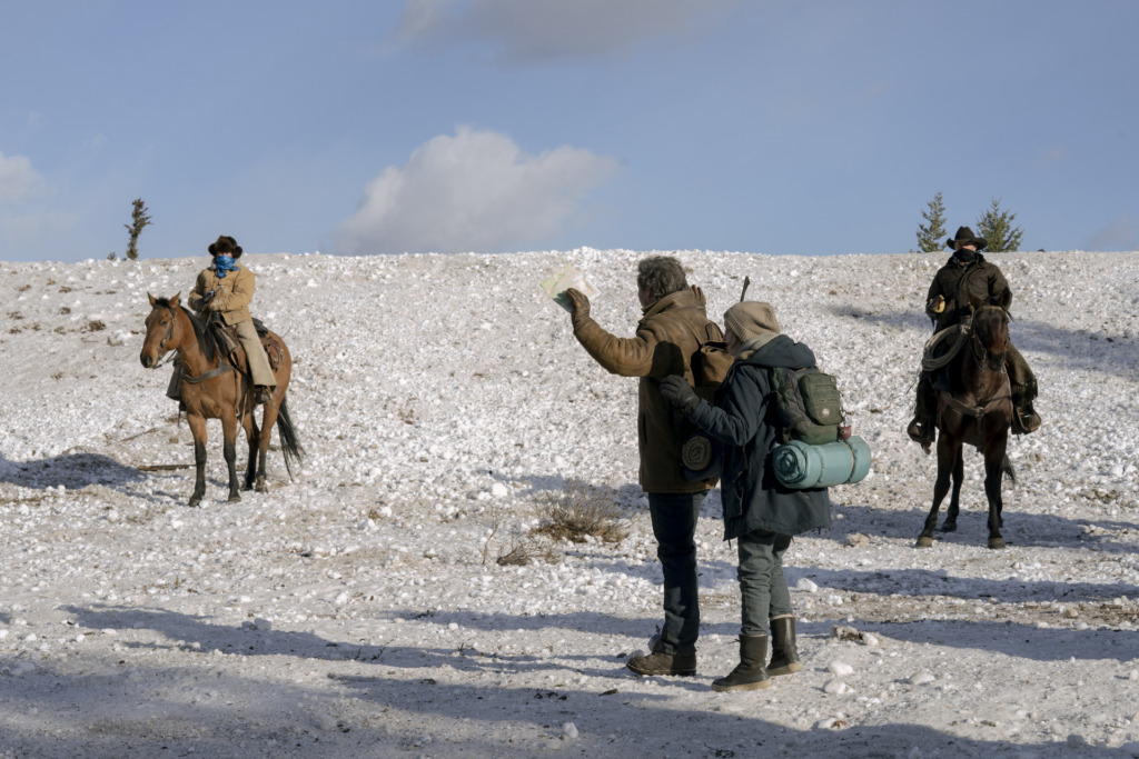 Joel (Pedro Pascal) and Ellie (Bella Ramsey) stand with their hands up as people on horseback surround them in the snow.