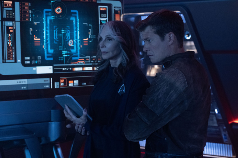 Gates McFadden as Dr. Beverly Crusher and Ed Speleers as Jack Crusher standing side by side with Beverly holding a data pad in her hand.