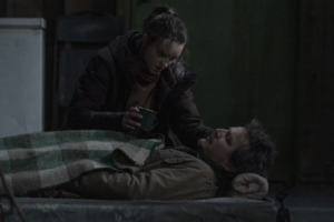 Ellie (Bella Ramsey) leans over an injured Joel (Pedro Pascal) with a cup of water.