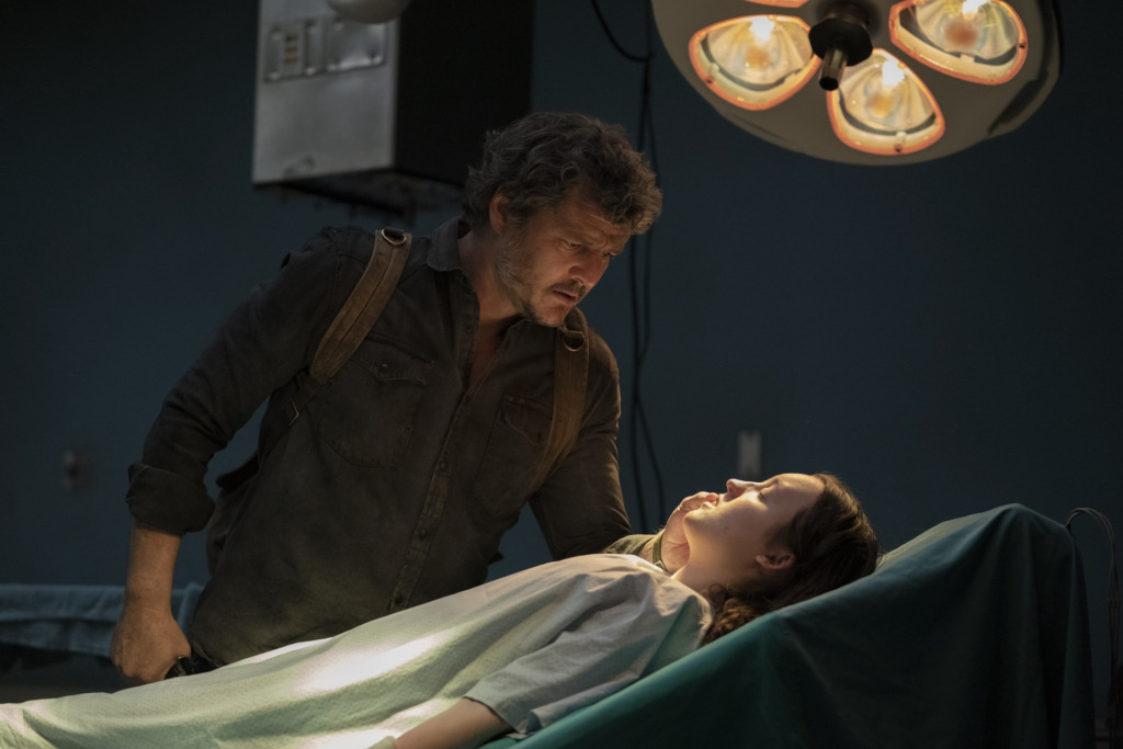 Joel (Pedro Pascal) leans over Ellie (Bella Ramsey), who is lying on a hospital table with a light shining on her.