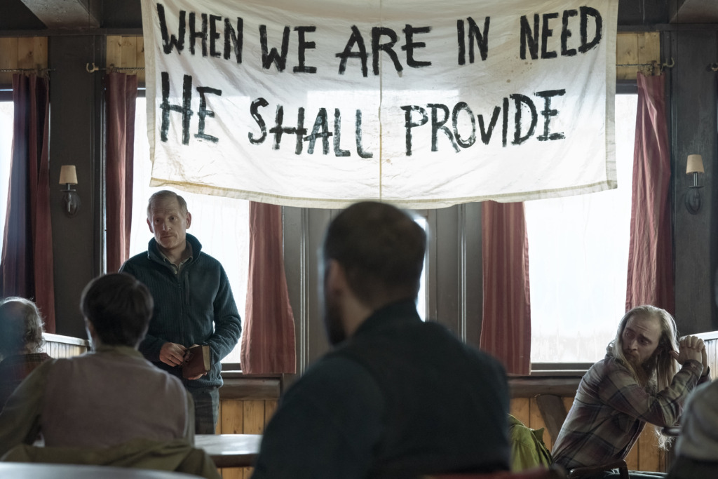 David (Scott Shepherd) stands in front of his group, holding a bible. Behind him, a banner hands, reading 'When we are in need, He shall provide'.