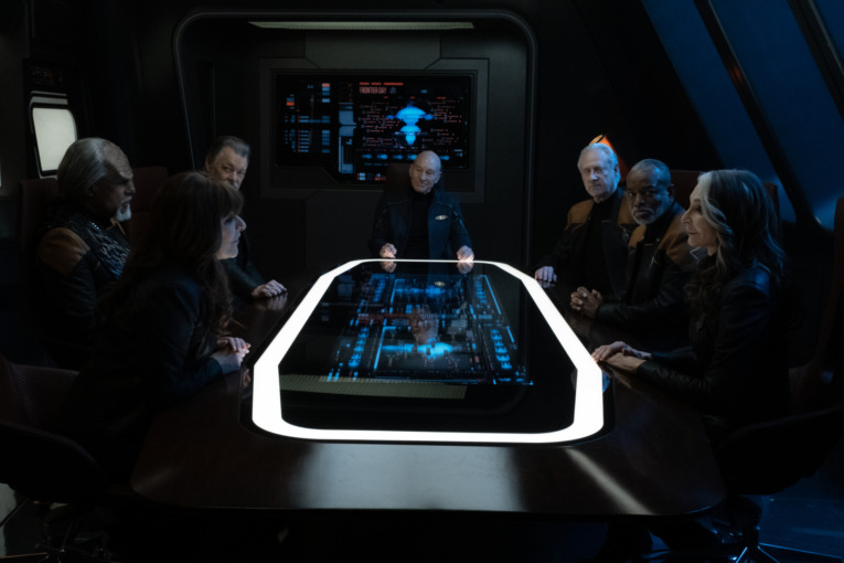 Jonathan Frakes as Will Riker, Brent Spiner as Data, Patrick Stewart as Picard, Michael Dorn as Worf, Marina Sirtis as Deanna Troi, LeVar Burton as Geordi La Forge and Gates McFadden as Dr. Beverly Crusher in "Surrender" Episode 308, Star Trek: Picard. The Crew sitting and gathered around a Conference Table with Star Trek themed graphic displays