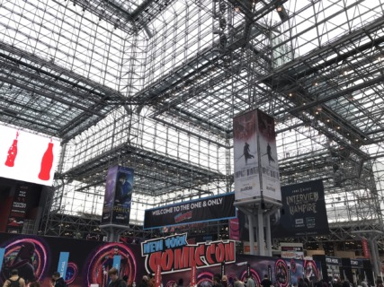 A photo of one of the entrances to NYCC inside the Javits convention center in 2022. This section of the building has a large glass ceiling with many support structures. Screens and ads are hung from these.