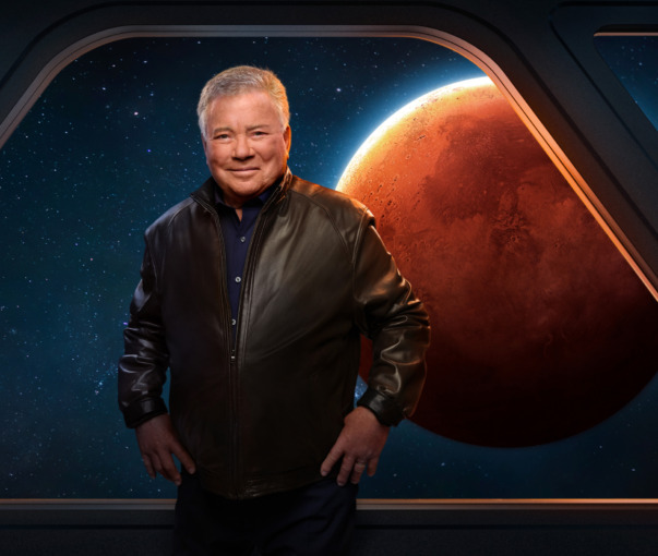 William Shatner standing and posing with hands on hips in front of a backdrop of a space window and the planet Mars in the background