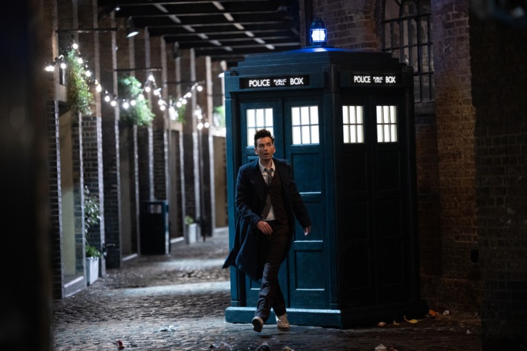 David Tennant as the 14th Doctor walks away from the TARDIS in a brick-paved alleyway