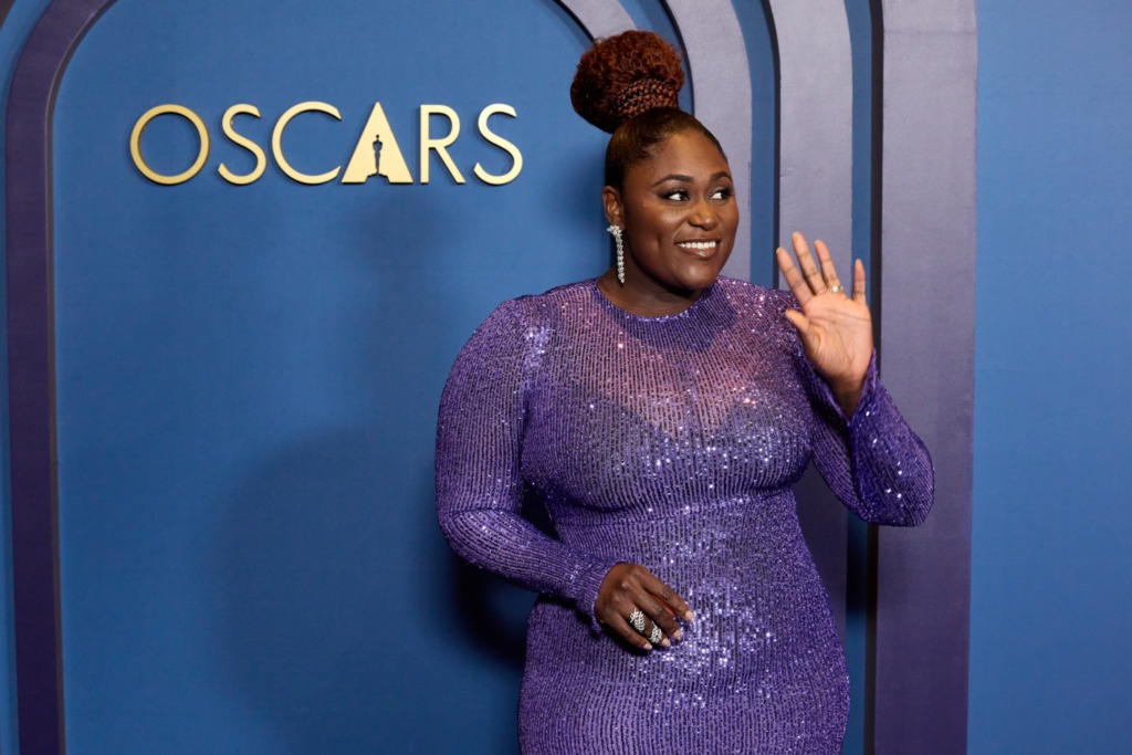 Danielle Brooks waving wearing a purple, sparkly dress.