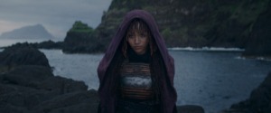 A young Black woman wearing a purple hooded robe stands in front of a body of water and rocky mountains.