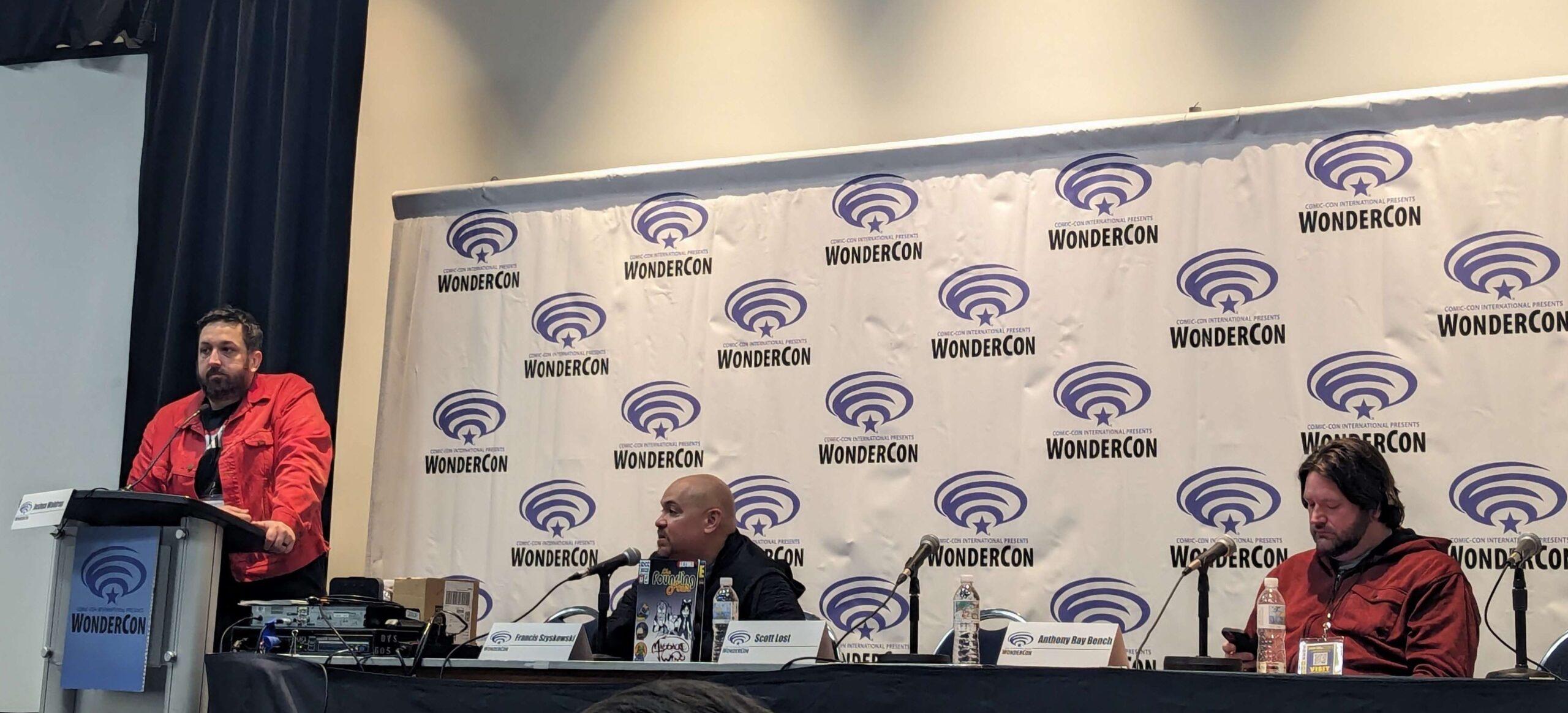 Ultima Comics panelists sitting at a long black booth with "wondercon" logo banner behind them.