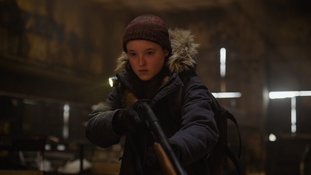 Ellie, in a winter coat and hat hold a shotgun down as she stares at something off camera.