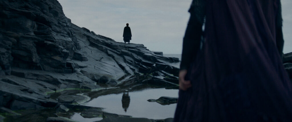 A dark figure stands on watery cliffs in the distance. Mae, turned away, faces toward the figure.