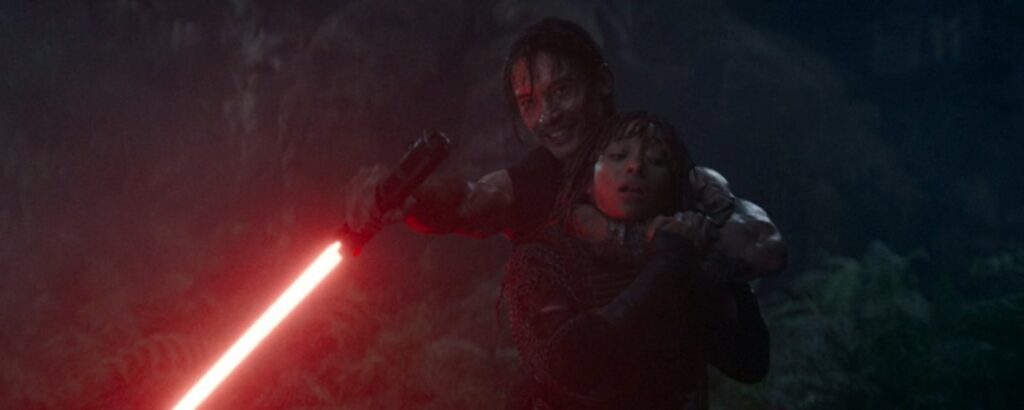 Qimir unmasked, holding Mae against his chest, with his red lightsaber held up in front of them.