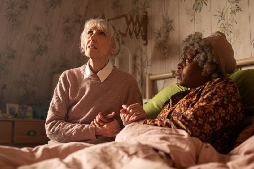 Mrs Flood (Anita Dobson) and Cherry (Angela Wynter) in Doctor Who's "Empire of Death" sit up in a bed, and hold hands, waiting in anticipation. 