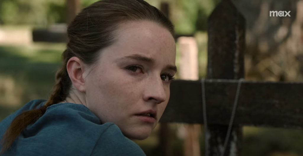 Abby, with tears in her eyes, looks away from the cross she's kneeling in front of.