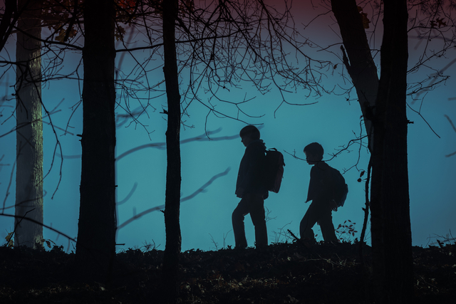 Two boys walking through woods are silhouetted against a light blue night sky.