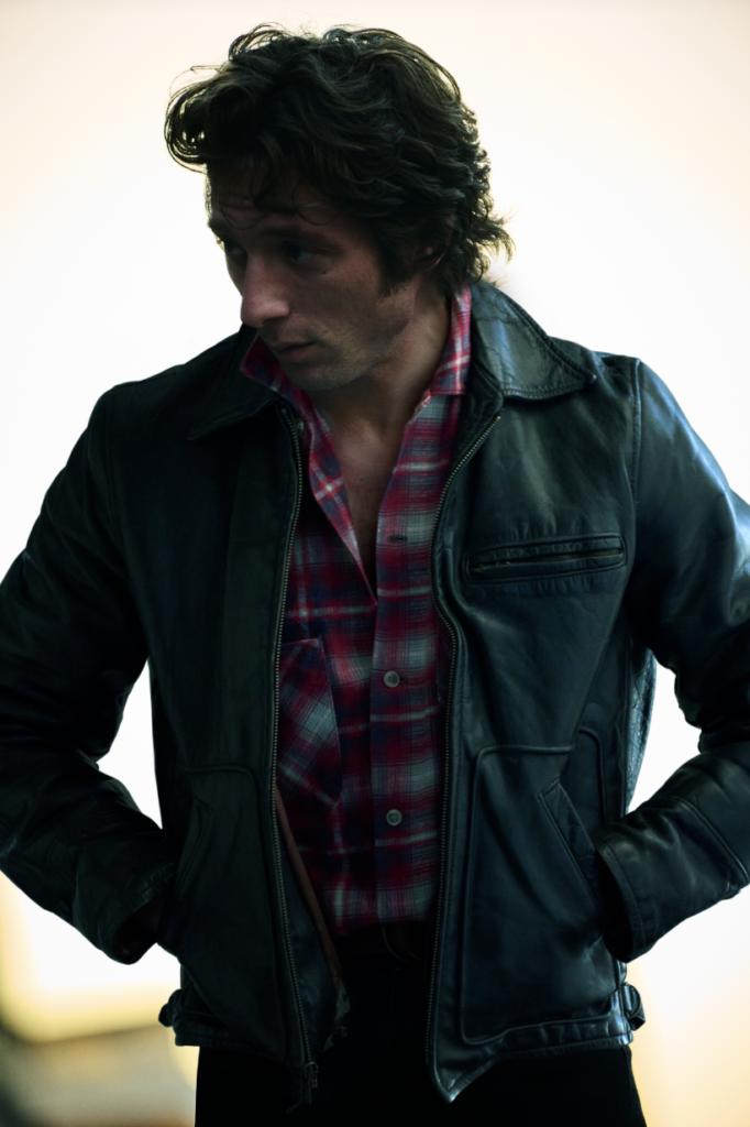 Jeremy Allen White as Bruce Springsteen in a black leather jacket over a red, blue and white flannel button-up shirt. White is looking off to the side with his hands in his jacket pockets.