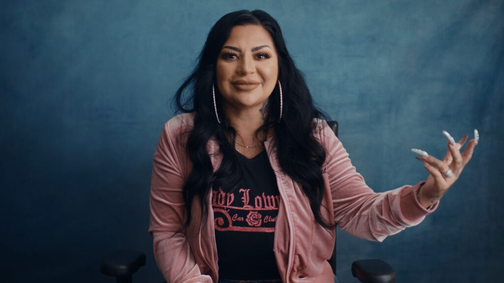 Sandy Avila From Lady Lowrider Club speaks directly to the camera. Sandy is wearing a pink jacket and a teeshirt to advertise her club