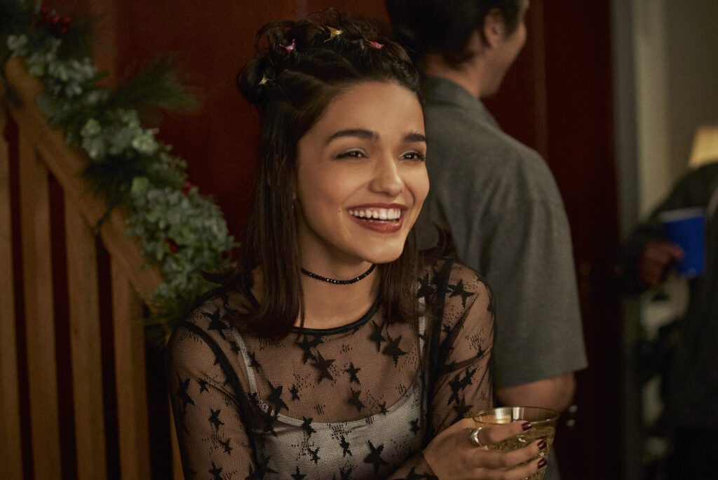 Rachel Zegler smiles and looks happy while at a party. She is wearing 90s era fashions, a lace see through shirt with stars and a visible white tank top. popular in the 1990s. 