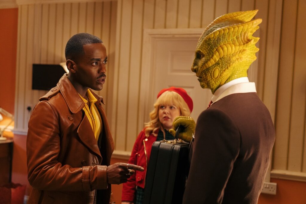 The Doctor (Ncuti Gatwa) speaks to the Silurian Time Hotel manager (Jonathan Aris)- a green-scaled reptile-like man- who is holding a briefcase. Joy (Nicola Couglan) looks in in the background. From "Joy to the World"