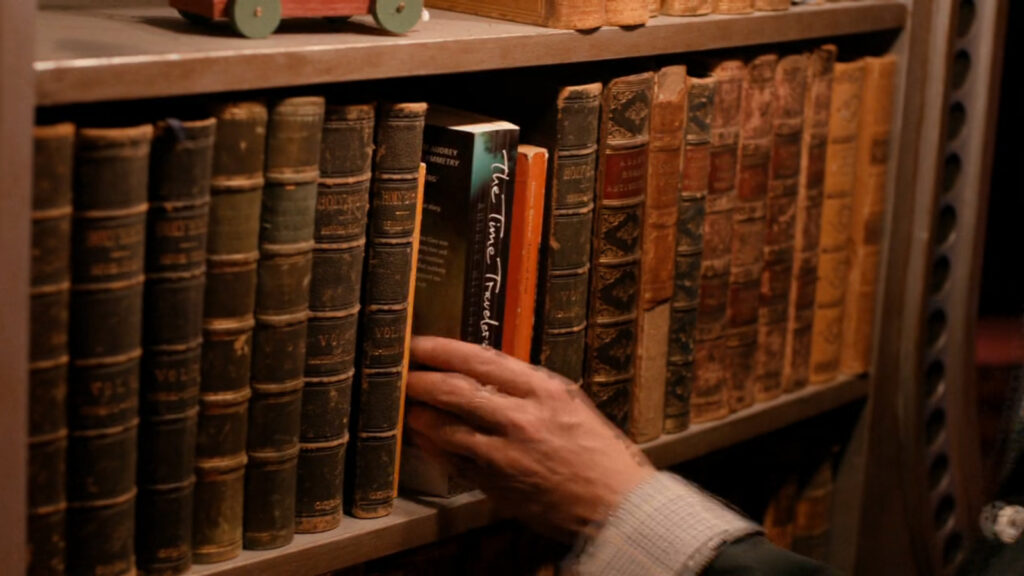 Clara's hand reaches for a copy of "The Time Traveler's Wife" on a bookshelf in the TARDIS