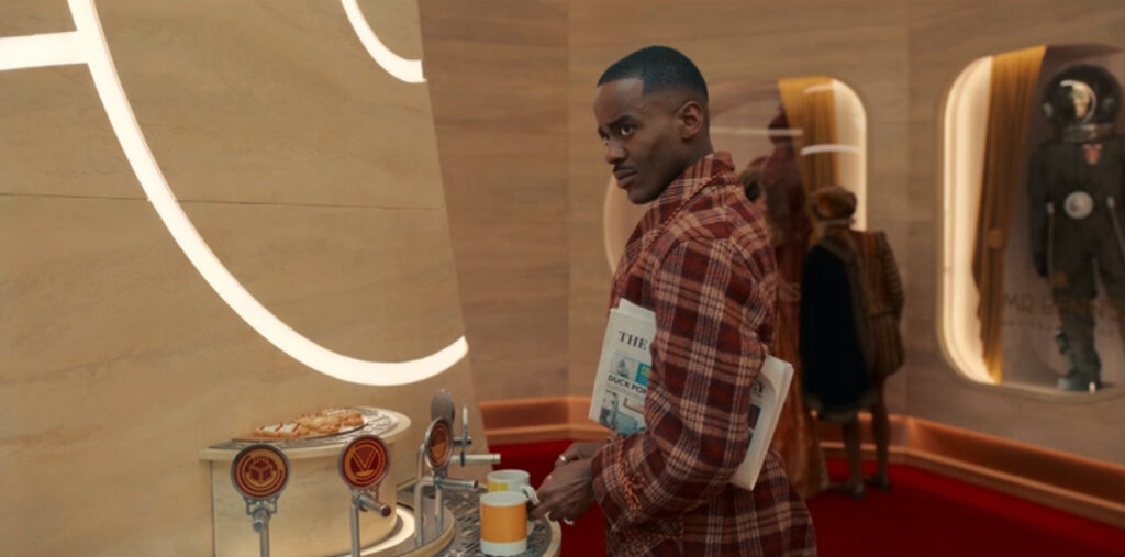 The Doctor in "Joy to the World" wearing a plaid robe with a newspaper tucked under his arm as he gets milk from a fancy dispenser in the Time Hotel lobby