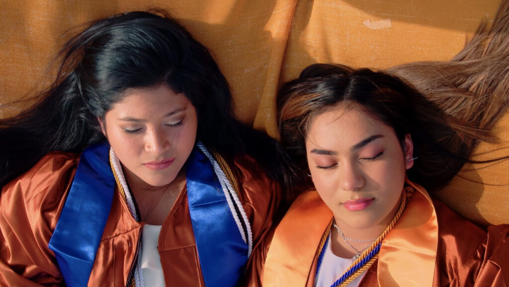 Two young Latina girls lie on a blanket in graduation robes, eyes closed against the sun.