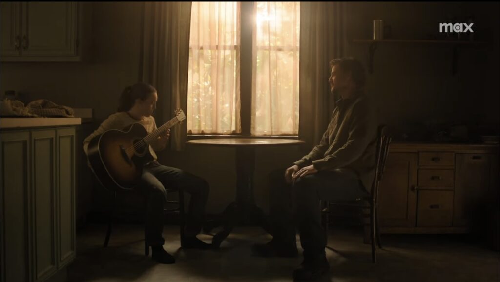 Ellie and Joel sit at a table. Joel watches as Ellie holds a guitar.