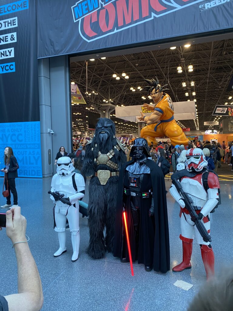 Star Wars cosplayers (a Stormtrooper, Black Krrsantan [a Wookie], Darth Vader, and a Stormtrooper with red accents on their armor) pose on the show floor at NYCC